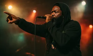 A man with dreadlocks in a black hoodie points forward while rapping into a microphone on a dimly lit stage.