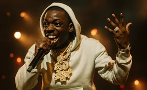 Rapper Bobby Shmurda on stage, wearing a hoodie and diamond chains, holding a microphone and gesturing.