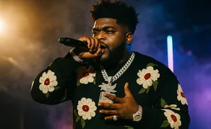 Rapper holding microphone, wearing a floral sweatshirt and diamond chains.