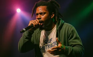 Rapper D.R.A.M. performs onstage with a microphone and wears a green hoodie over a t-shirt with 