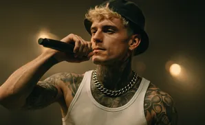Man with blond hair, tattoos, and a chain necklace sings into a microphone.