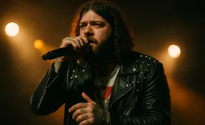 A tattooed man with long, dark hair sings into a microphone while wearing a studded leather jacket.