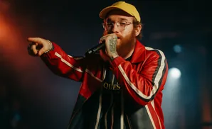 Man with red beard, glasses, and yellow cap sings into a microphone, wearing a red, white, and blue jacket.