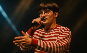 A man with glasses and a mustache sings into a microphone, wearing a red and white striped sweater.
