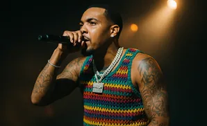 A man in a colorful crocheted tank top and chains sings into a microphone on stage.