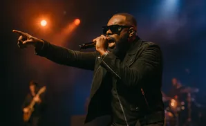 A Black man in sunglasses and a black jacket sings into a microphone, pointing with his left hand on a dimly lit stage.