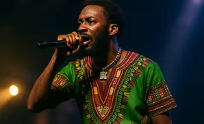 A Black man in a green dashiki sings into a microphone on a dark stage with blue and orange lighting.