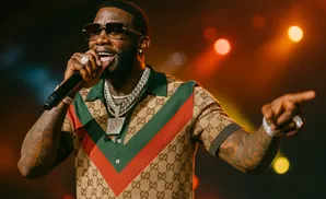 Rapper Gucci Mane on stage wearing a Gucci shirt, sunglasses, and jewelry, holding a microphone.