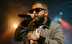 A man with a beard and sunglasses speaks into a microphone, wearing a grey jacket.