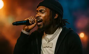 A rapper in a beanie and chains performs into a microphone, with warm stage lighting.