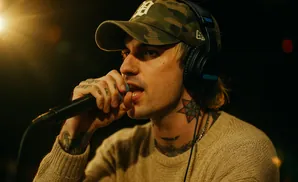 A man in a camouflage hat and headphones sings into a microphone, with tattoos visible on his hand and neck.