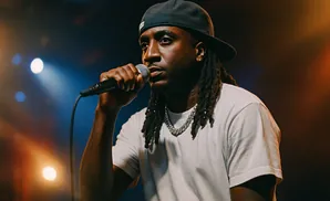 A Black man wearing a black hoodie and baseball cap sings into a microphone under stage lights.