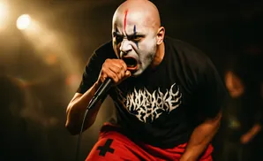 A bald man with face paint sings into a microphone on a dimly lit stage.