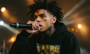 A rapper with dreadlocks and tattoos performs into a microphone on stage, illuminated by warm stage lights.