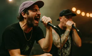 A man in a purple cap sings into a microphone, eyes closed, mouth wide open.
