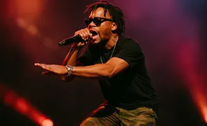 A male rapper performs on stage, wearing sunglasses and holding a microphone.
