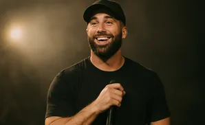 A man in a black t-shirt and baseball cap smiles while holding a microphone.