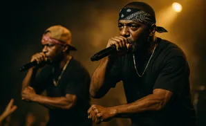 Two men rapping into microphones on a dimly lit stage, one in a black bandana, the other in a tan baseball cap.