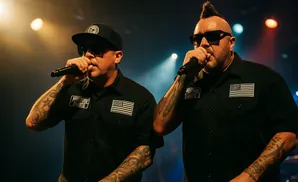 Two men with tattoos on their arms wearing black shirts and sunglasses, rapping into microphones on stage.