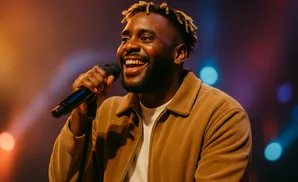 A Black man with dreadlocks smiles and sings into a microphone. He wears a brown corduroy jacket.