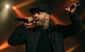 A tattooed man in a black baseball cap and jacket sings into a microphone on stage, illuminated by orange spotlights.