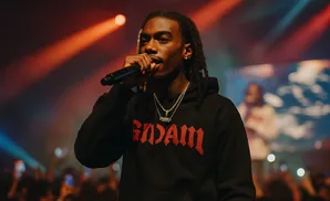 Rapper performing on stage, holding a microphone, wearing a black hoodie with red text.