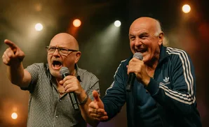 Two bald men singing into microphones on stage, one pointing.