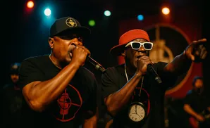 Two men in black t-shirts rap into microphones on a dimly lit stage.