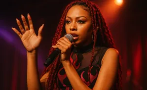 A singer with red dreadlocks holds a microphone and raises her hand on a dimly lit stage.