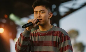 A young Asian man in a striped sweater holds a microphone and sings on stage.