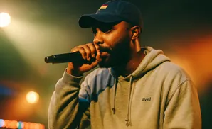 A man wearing a grey hoodie and a blue baseball cap with a flag on it, holding a microphone to his mouth while performing on stage.