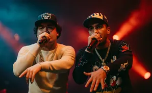 Two rappers perform on stage, holding microphones and wearing baseball caps and gold chains.