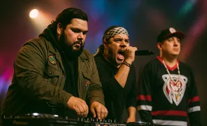Three men perform on stage, one DJing and two singing into microphones, with colorful stage lights in the background.