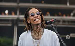 Rapper Wiz Khalifa smiles and performs into a microphone, wearing sunglasses and a white t-shirt with numerous necklaces.