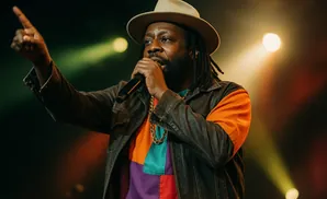 A Black man in a hat and jacket sings into a microphone, pointing his finger upwards.