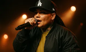 A woman wearing a black baseball cap and jacket performs into a microphone under warm stage lights.