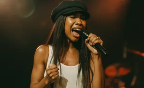 A Black woman in a white tank top and black cap sings into a microphone on a dimly lit stage.