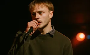 A young man with blonde hair holds a microphone to his mouth, wearing a dark green sweater over a collared shirt.