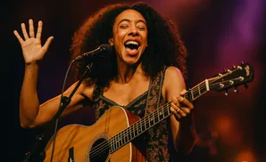 A woman with dark, curly hair sings into a microphone while playing an acoustic guitar and holding her left hand up.