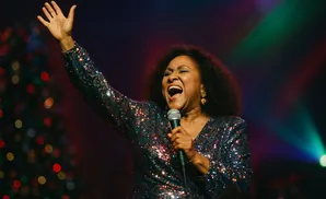 Singer in a sequined dress performs on stage, holding a microphone with her arm raised.