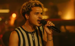 A Black man with curly hair sings into a microphone while wearing a striped tank top.