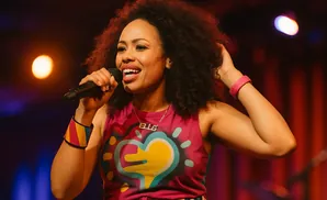Woman singing into a microphone, wearing a colorful shirt and jewelry.