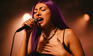 Singer with purple hair sings into a microphone, eyes closed, with a hand on her chest.
