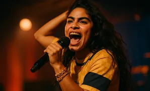A woman with dark curly hair sings into a microphone, her mouth wide open in song.