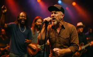 A band performs on stage with red and blue lights. The lead singer, wearing a baseball cap and brown shirt, sings into a microphone.