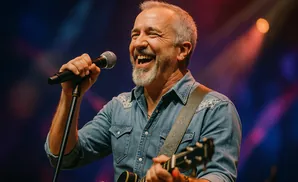 A smiling man with gray hair and a beard sings into a microphone while playing a guitar.