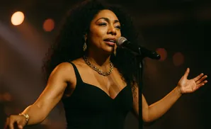 Singer with curly hair wearing a black dress and gold necklace performs into a microphone.