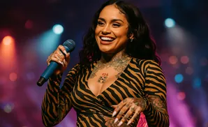 A smiling Black woman with tattoos holds a blue microphone while performing on a stage with colorful lighting.