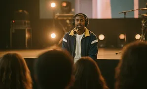 Man wearing headphones and a blue jacket sings into a microphone on stage, with audience in foreground.