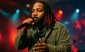 A man with dreadlocks and red accents raps into a microphone on a stage with colorful lights.